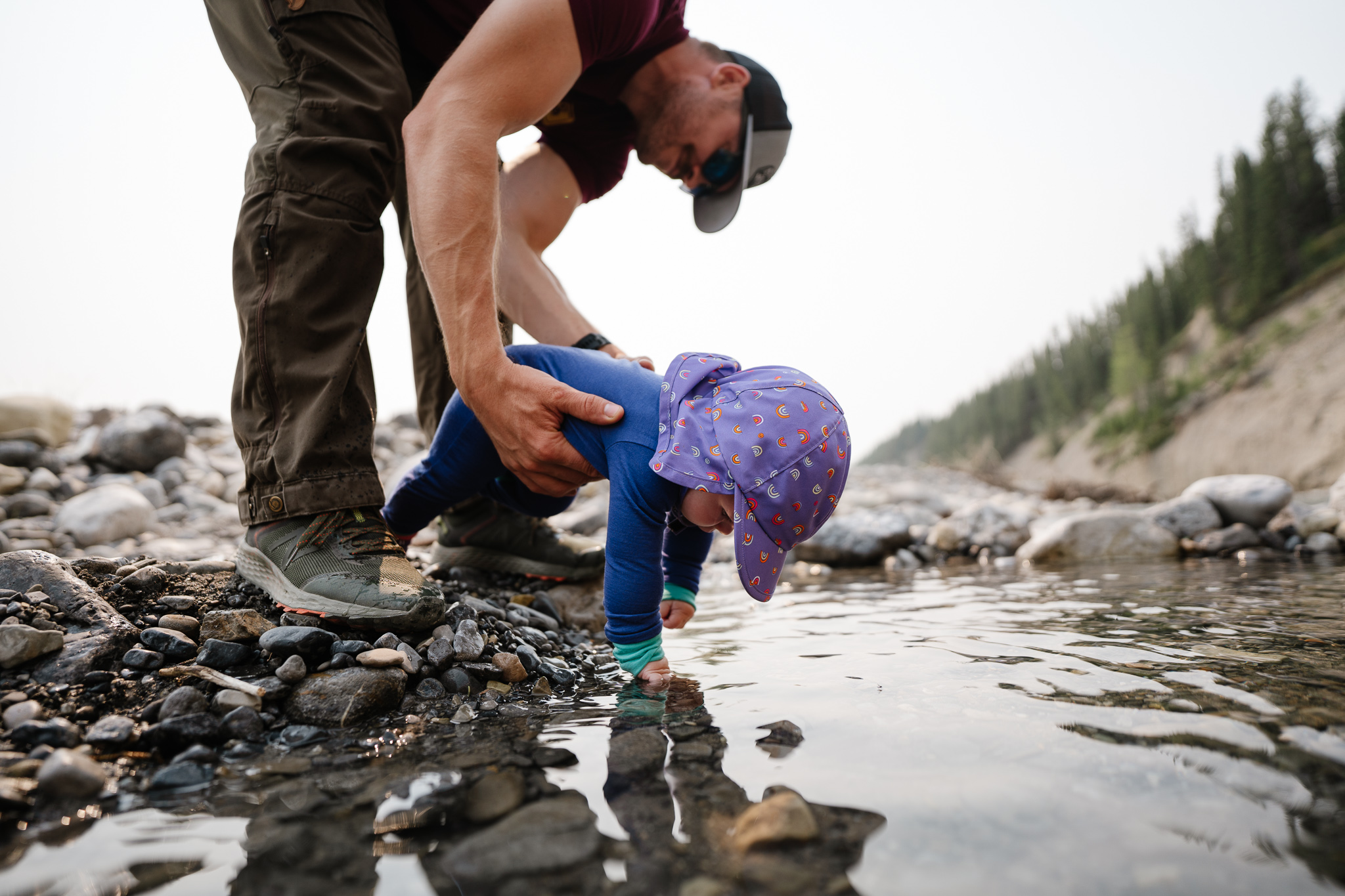 dads holds baby while she reaches into stream to play with rocks while camping