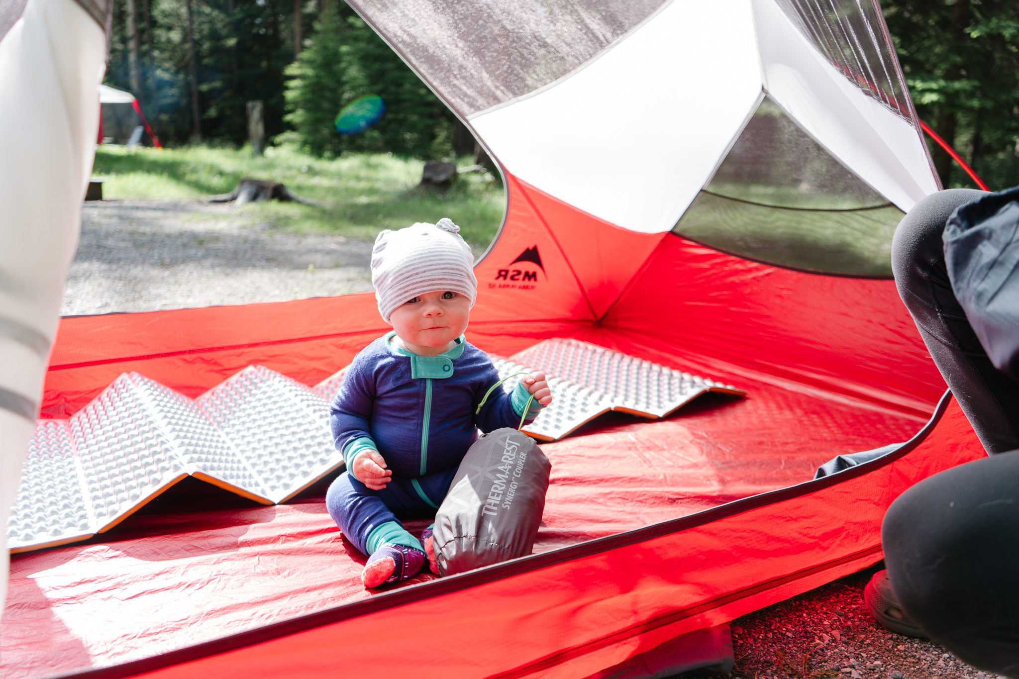 Baby sits inside tent holding a sleeping pad while unpacking for camping