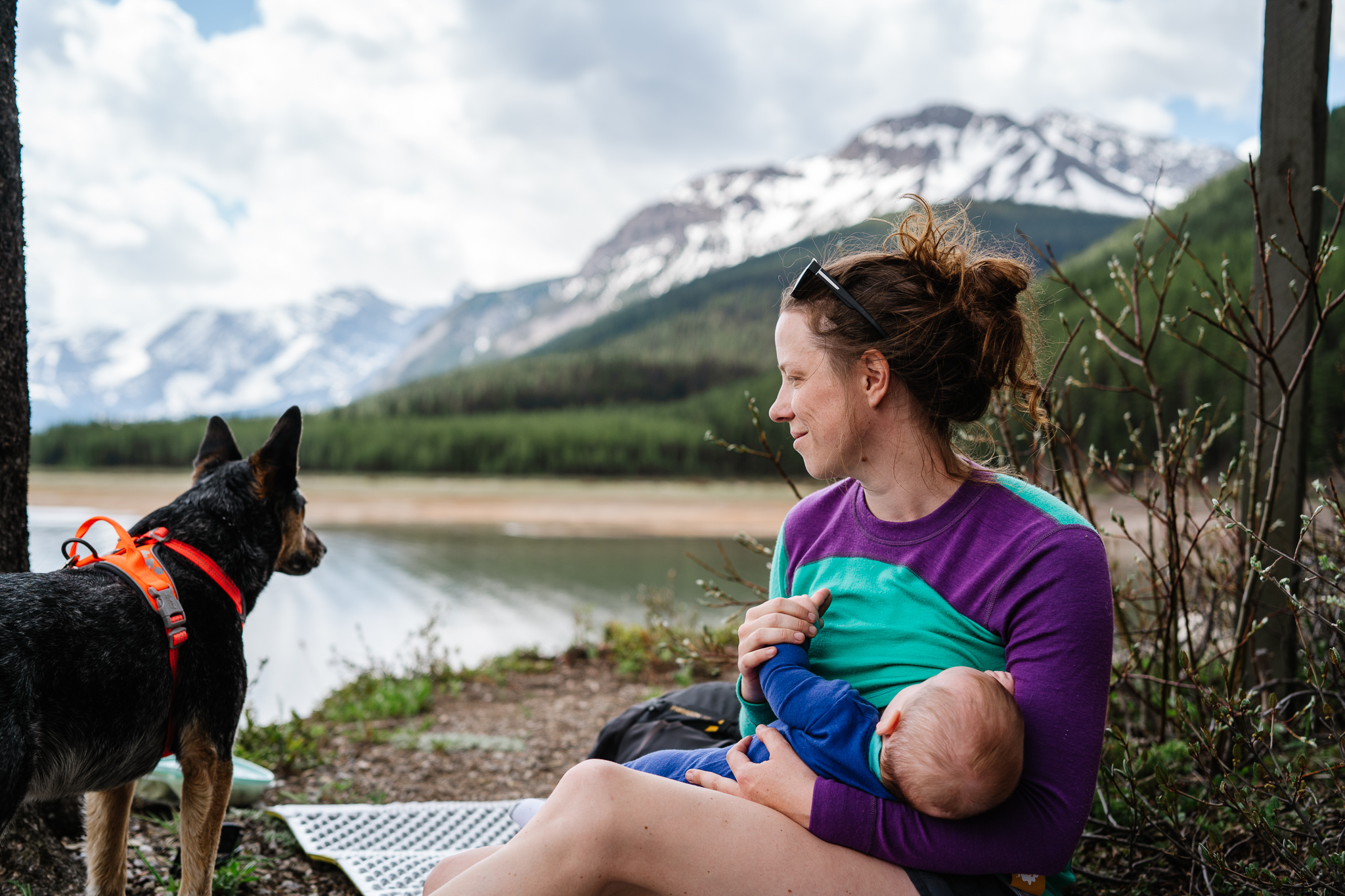 Mom sits lakeside in the mountains breastfeeding her baby while she looks at her cattle dog standing next to her. 