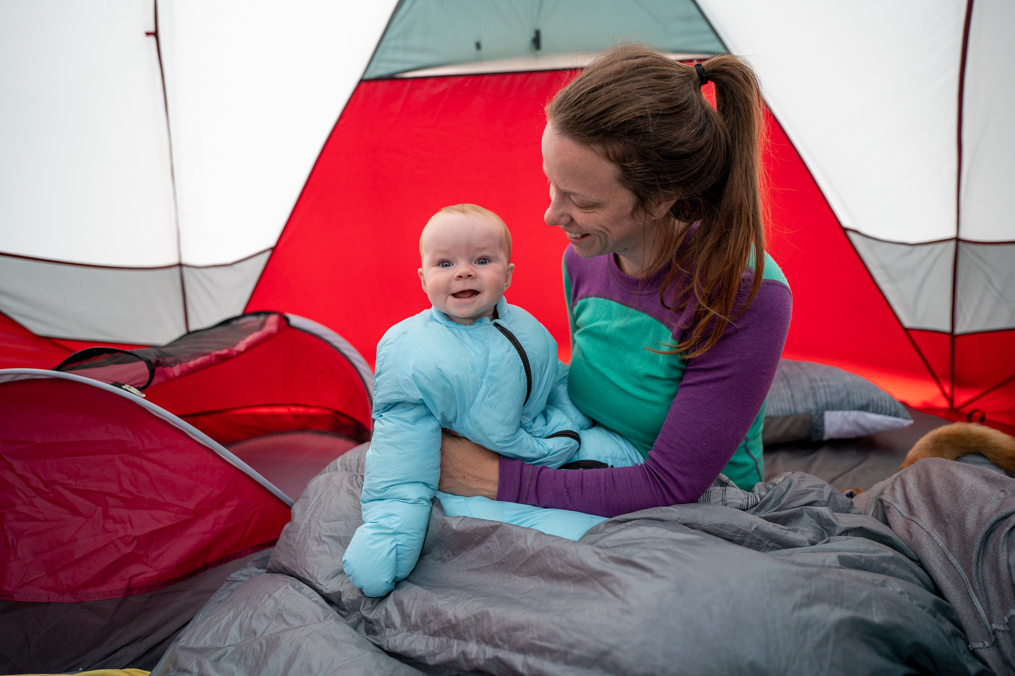 Mom in merino wool breastfeeding top smiles at baby wearing an infant sleeping bag while camping in Alberta.
