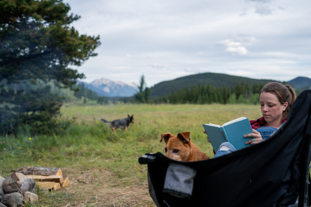 Girl sits in a camping chair with a red dog reading Untamed while black dog plays in the grass behind them. 