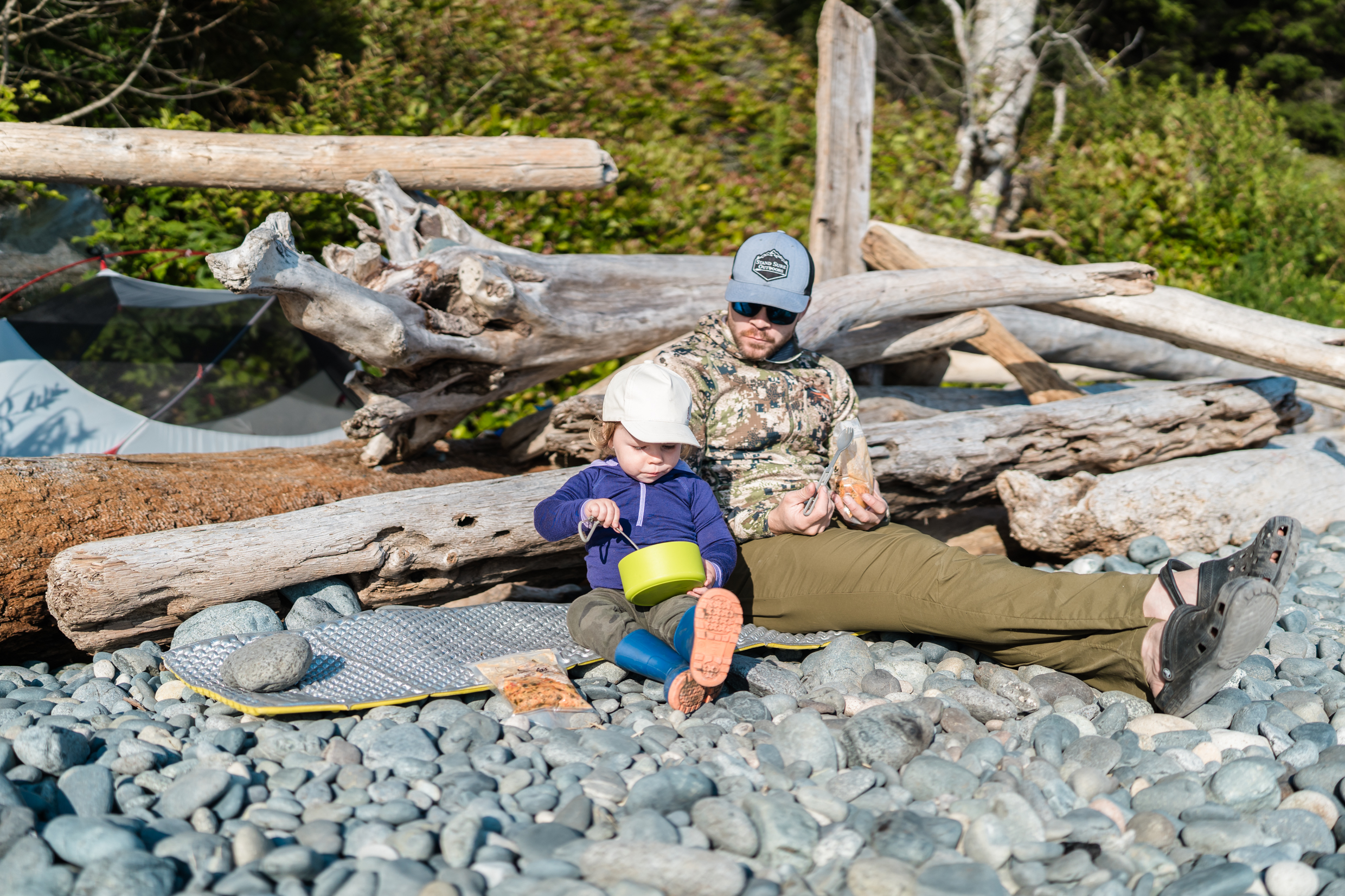 Dad and daughter sit on a beach together while backpacking eating dehydrated meals for dinner. 
