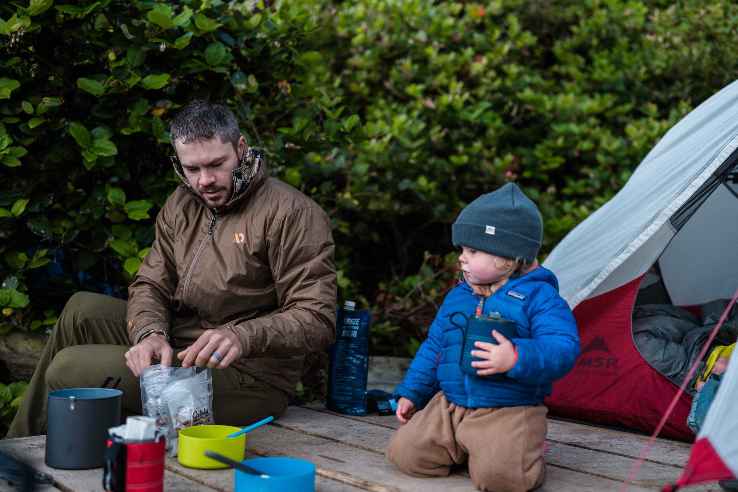 Man in brown jacket sits on tent platform preparing breakfast while backpacking with his toddler who sits beside him.