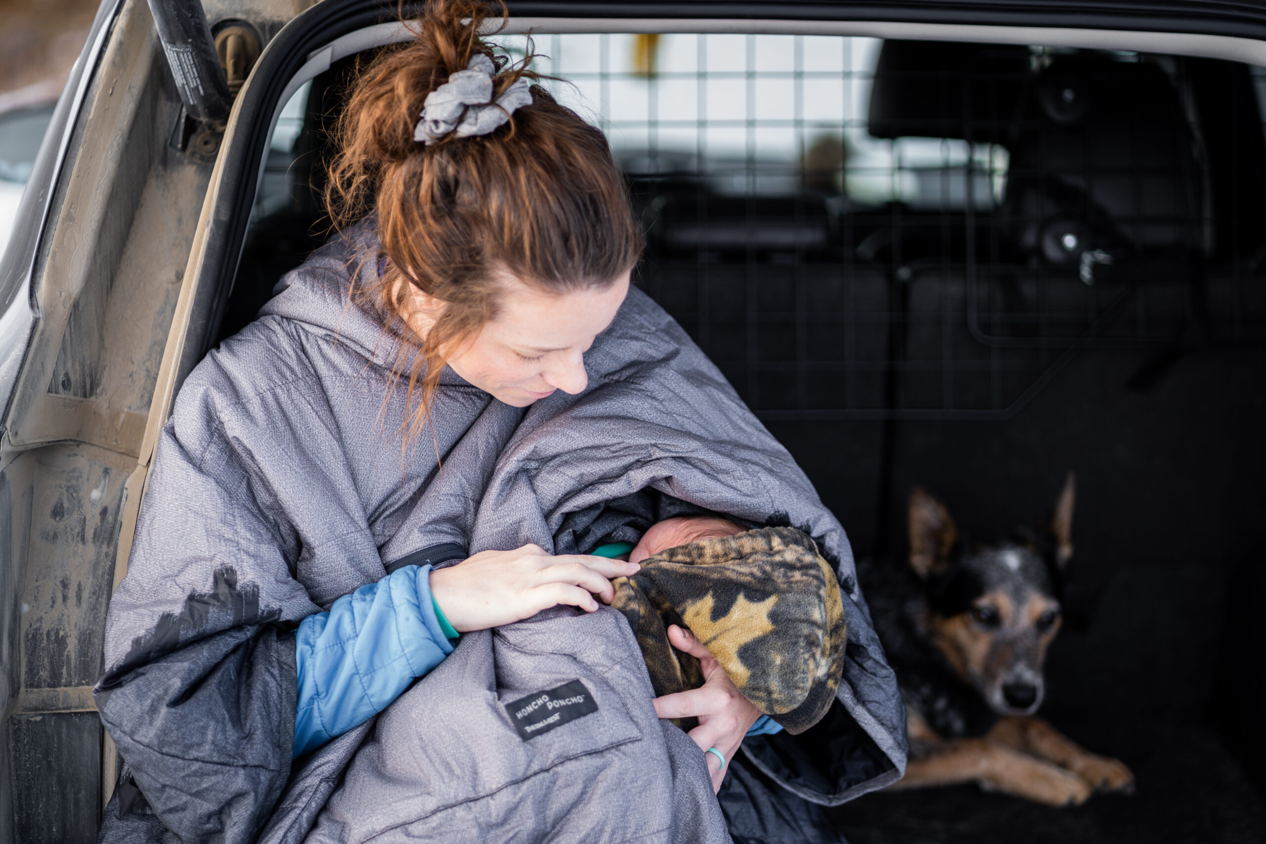 woman wearing a honcho poncho breastfeeds her baby during the winter in the back of the vehicle with her dog beside her before heading out hiking with babies