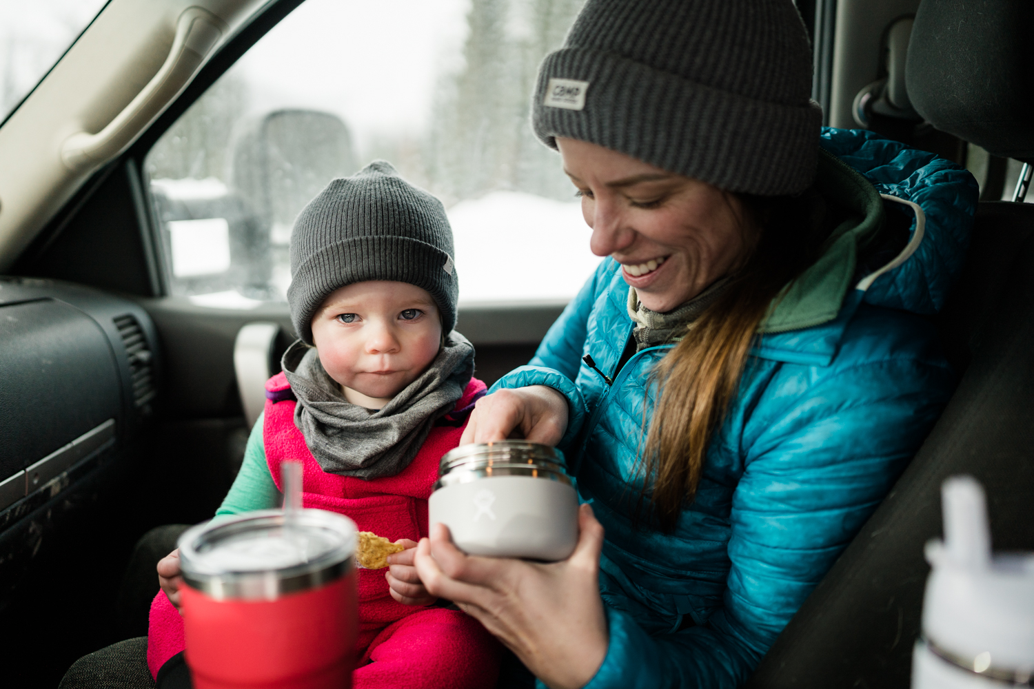 baby wearing pink fleece overalls with a grey toque and grey neck warmer sist on moms lap in truck after a winter hike eating out of insulated food container 