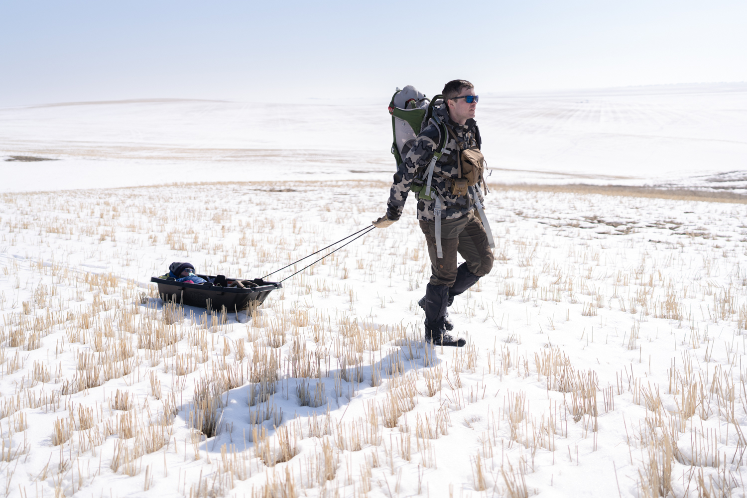 dad wearing camo pulls sleeping toddler in a black sled along snowy ground in a field 
