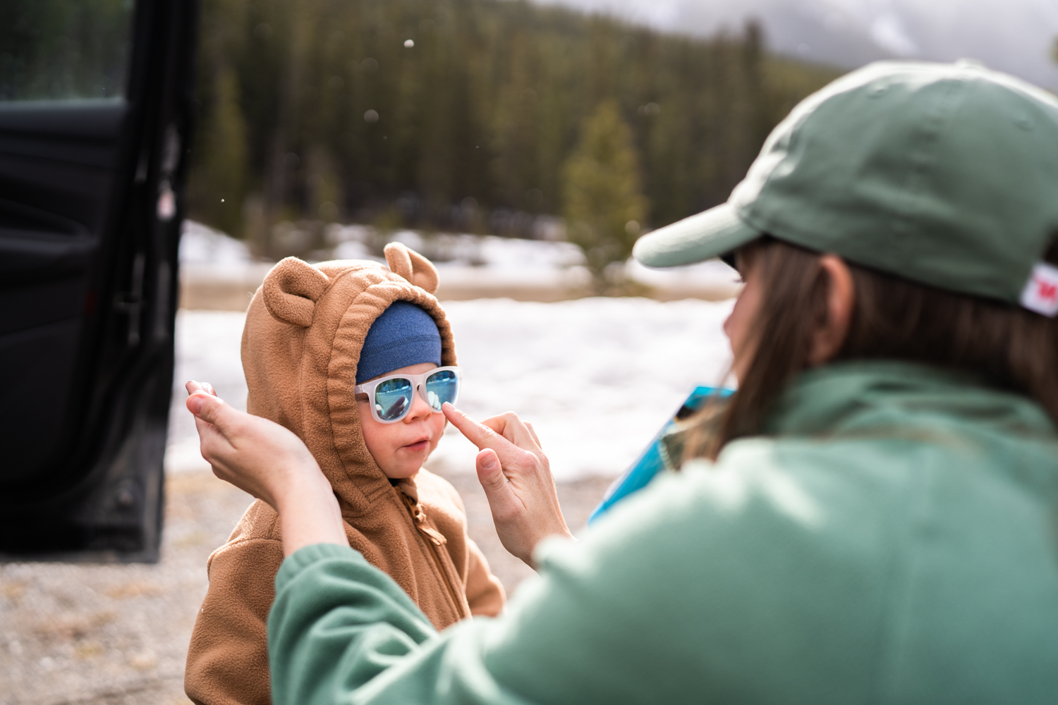 mom in green fleece and green hat applies sunscreen to toddlers face while she wears a hat, sunglasses, and a brown bear fleece before heading off on a hike