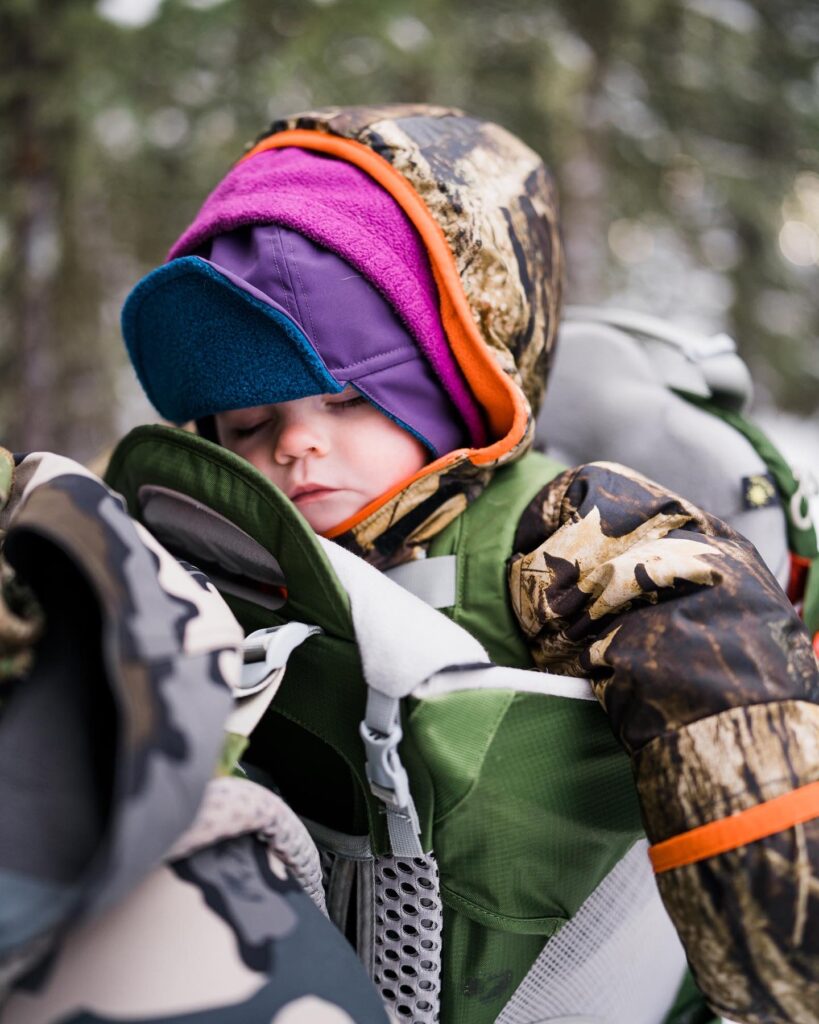 baby sleeps in a green backpack carrier while winter hiking and wearing many layers to stay warm