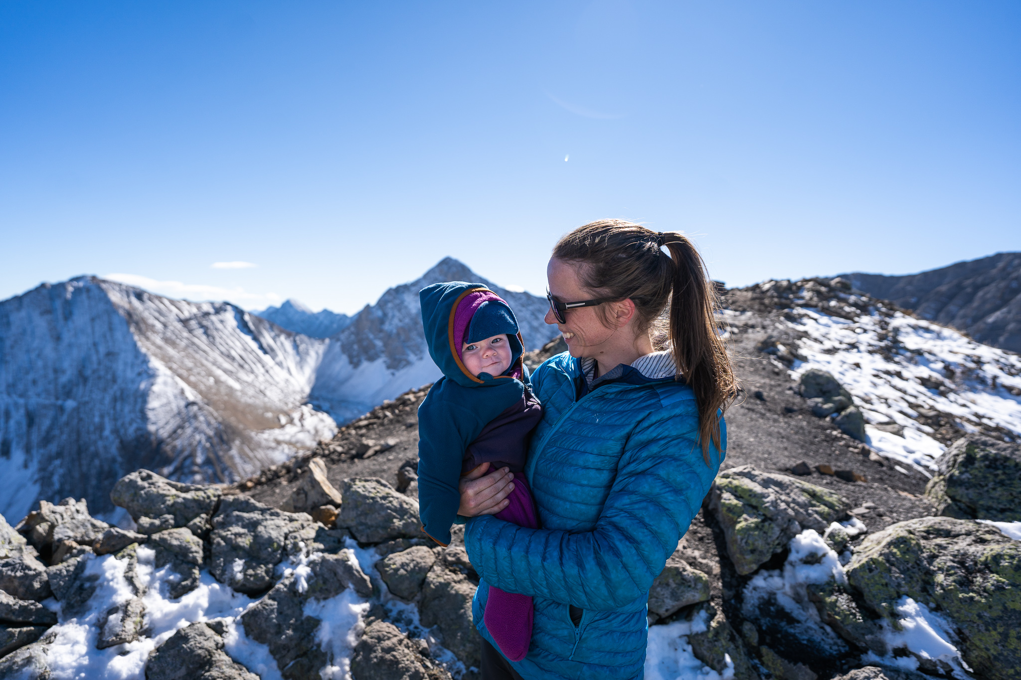 baby wearing fleece layers smiles at camera while being held by mom standing up top of a snowy ridge while hiking 