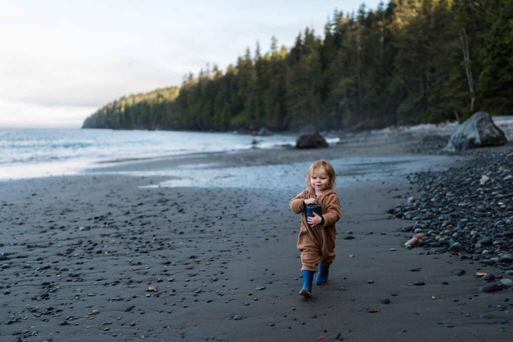 toddler walks along a beach at sunrise with blue rainboots and a blue GSI mug
