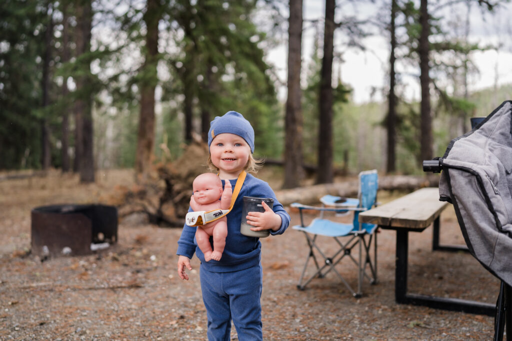 toddler in blue PJs holds cup of milk and has a baby tucked into a headlamp around her neck while car camping
