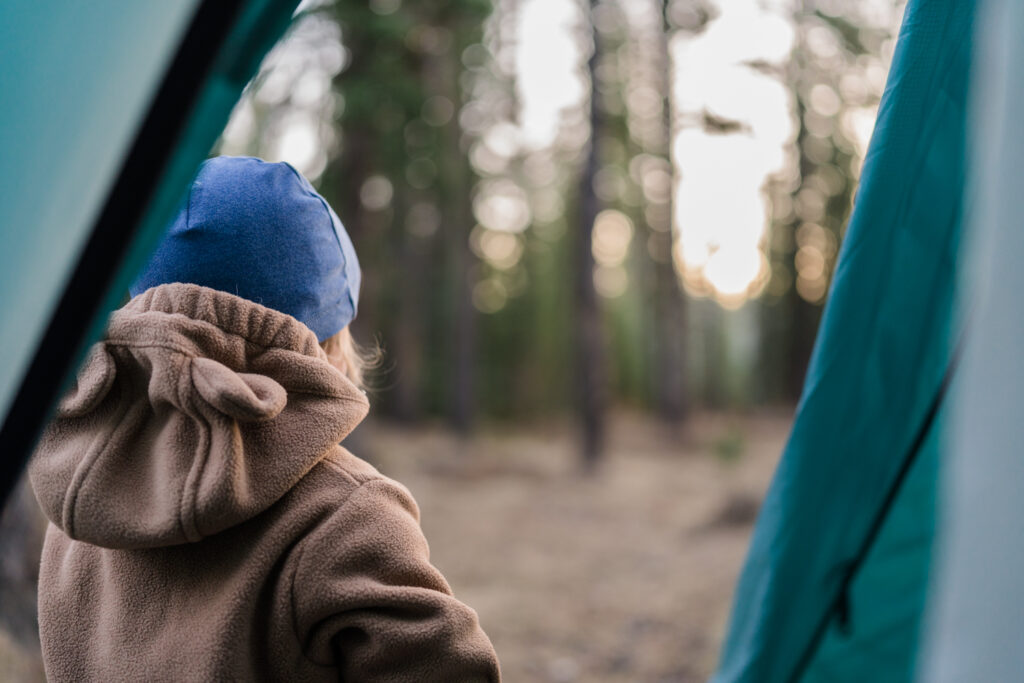 toddler in fleece suit and blue hat looks out at the sun coming through the trees while standing outside of a blue tent while camping